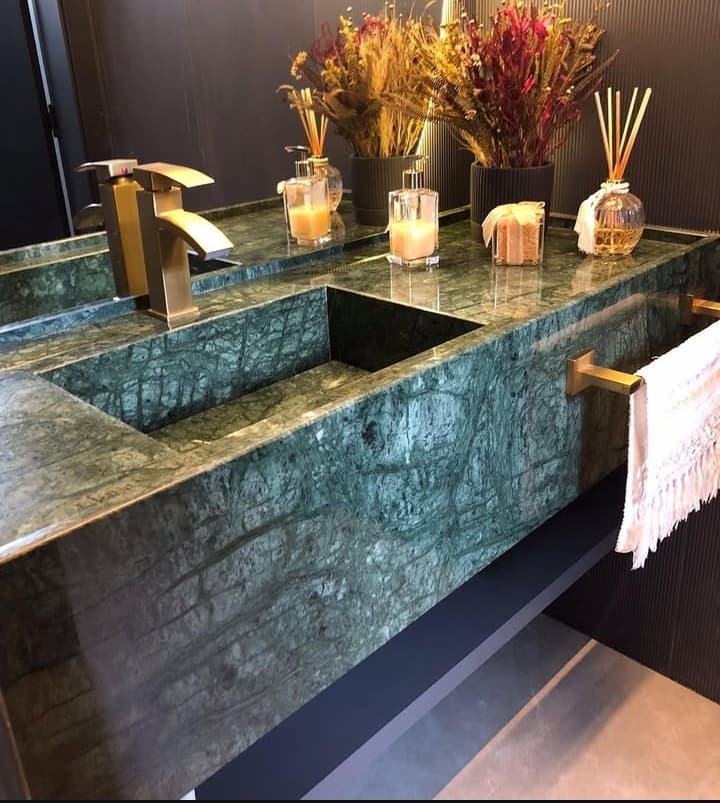 Bancada de banheiro em mármore verde com cuba esculpida e metais em tom de ouro velho.
