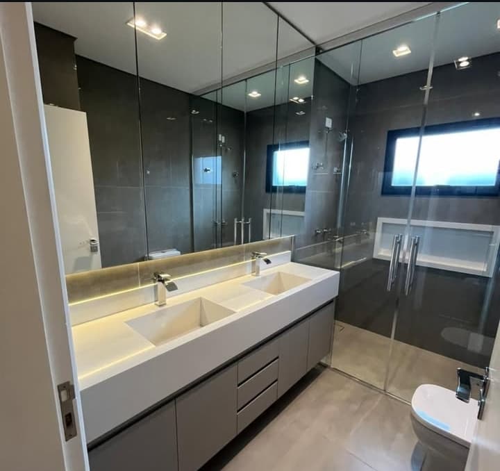 Banheiro amplo e moderno com bancada dupla e cuba esculpida em quartzo branco, armários cinza e espelho grande com iluminação embutida.